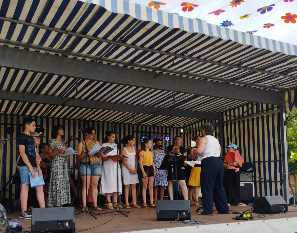 Marina Graf, de dos, portant un débardeur blanc et un pantalon foncé, dirige une chorale sur la même scène extérieure couverte par un chapiteau rayé bleu et blanc. Les choristes, enfants et adultes, tiennent des partitions et se tiennent en ligne derrière les micros. La scène est décorée de guirlandes colorées en forme de fleurs suspendues au plafond.