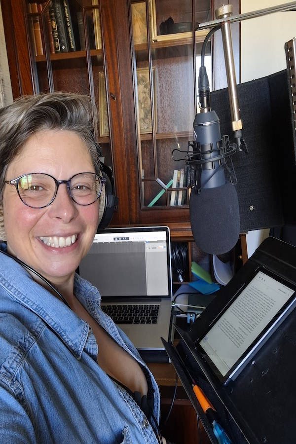 Marina Graf sourit devant son micro de studio. Elle porte un casque audio et des lunettes, avec un ordinateur portable et une tablette affichant un texte. Elle est installée dans un espace de travail avec bibliothèque en arrière-plan.