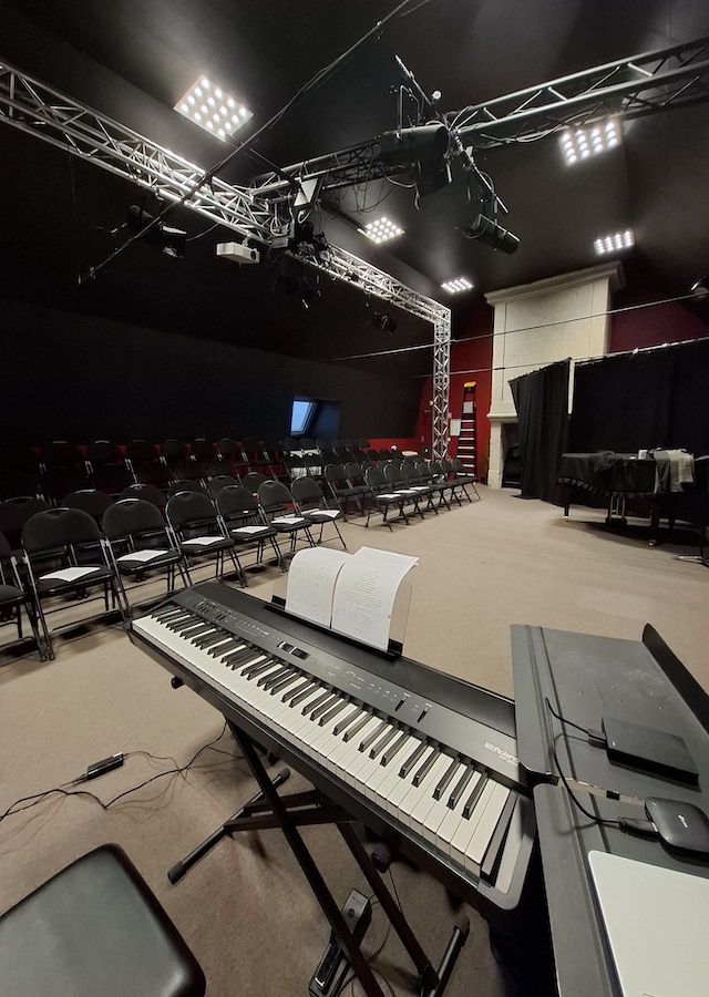 Salle de spectacle avec des rangées de chaises vides face à un clavier numérique Roland posé sur un pied, avec une partition ouverte. Un grand dispositif lumineux et technique suspendu au plafond éclaire la pièce.