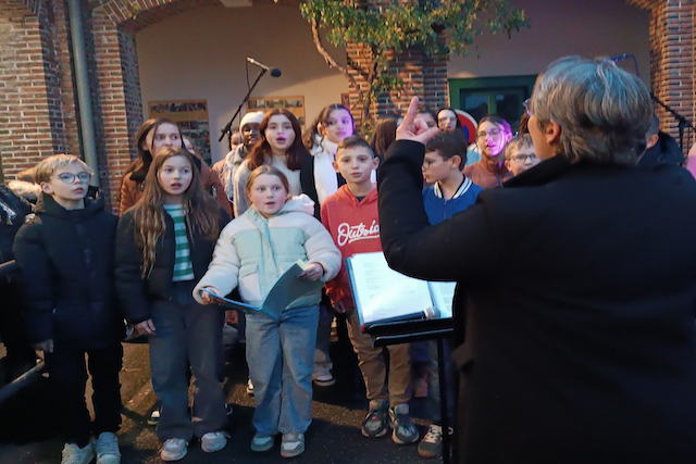 Marina Graf, cheffe de chœur, vue de dos, dirige un groupe d’enfants et d’adolescents qui chantent en extérieur, devant un mur de briques. Elle lève la main pour donner les indications rythmiques et vocales.