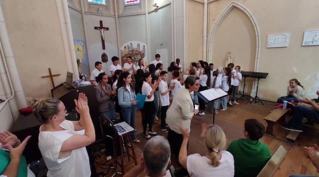Marina Graf, en veste claire, dirige un groupe d’enfants vêtus de t-shirts blancs, rassemblés devant l’autel d’une église. Le public applaudit, et la scène dégage une ambiance festive et collective.