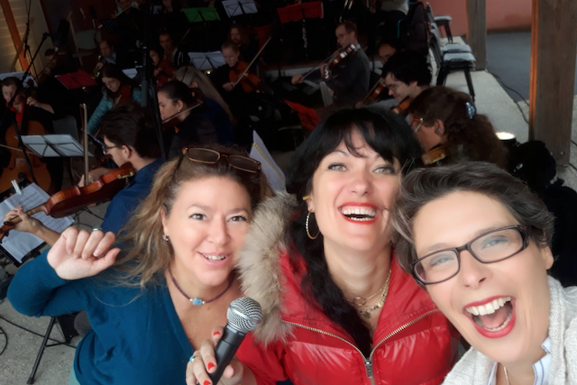 Marina Graf à droite sourit largement avec deux amies, l’une tenant un micro, devant un orchestre en répétition avec des violonistes et d’autres musiciens à cordes jouant à l’arrière-plan. Les trois femmes posent joyeusement en gros plan.