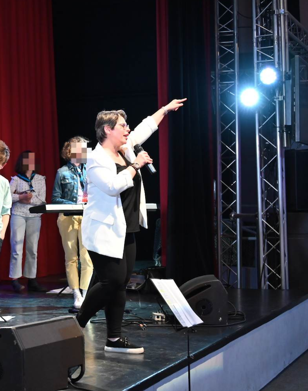 Marina Graf, vêtue d’une veste blanche et d’un haut noir, chante et dirige sur scène un groupe de jeunes choristes. Elle tient un micro d’une main et lève l’autre bras en direction du public. Derrière elle, on distingue un clavier et des lumières de spectacle.