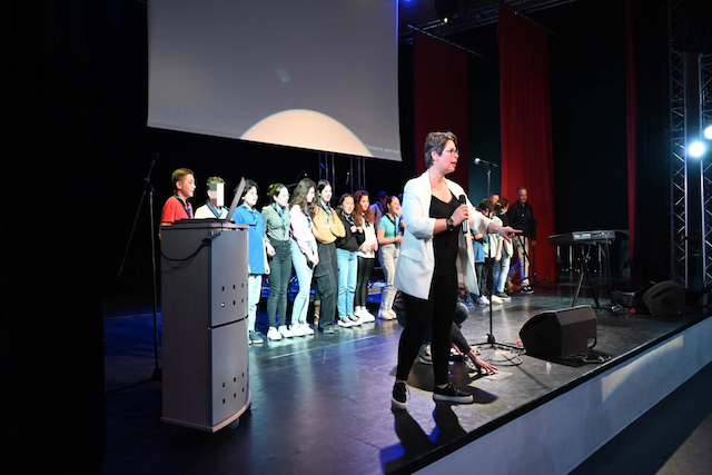 Marina Graf, cheffe de chœur, sur scène devant un groupe d’élèves qui chantent en chorale. Elle porte une veste claire et dirige avec énergie, un micro devant elle. La scène est éclairée, avec rideaux rouges et projecteurs, créant une ambiance de spectacle.