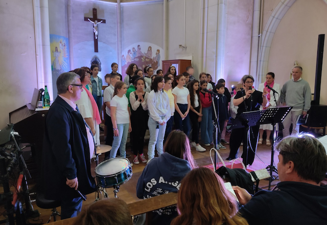 Dans une église, Marina Graf dirige et chante avec un micro devant une chorale d’enfants et d’adolescents vêtus de blanc et de couleurs claires. Elle est à droite de l’image, habillée en noir et blanc, devant des pupitres et un tambourin posé. Le public est visible au premier plan.