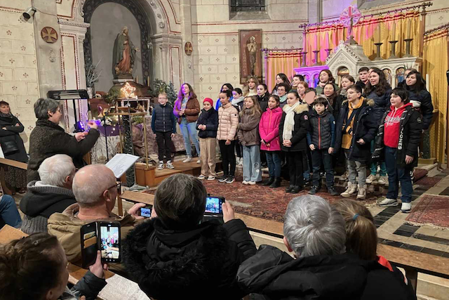 Marina Graf, de dos, dirige une chorale d’enfants vêtus de manteaux d’hiver dans une église décorée d’un autel et de tentures dorées. Le public prend des photos et des vidéos avec ses téléphones, assis sur les bancs de l’église.