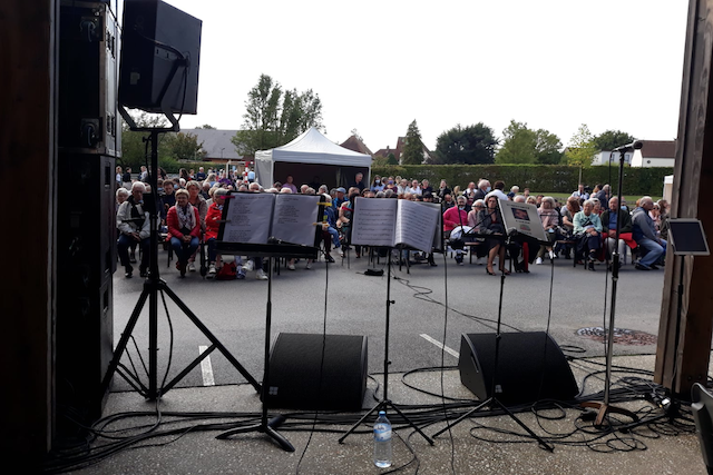 Vue depuis une scène extérieure d’un concert. On voit plusieurs pupitres avec partitions, micros et enceintes au premier plan, et en arrière-plan un public assis sous des tentes et à l’air libre.