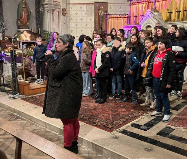Marina Graf, micro à la main, s’adresse à un public pendant qu’un groupe d’enfants chante sur scène dans une église décorée avec un autel et des rideaux colorés.