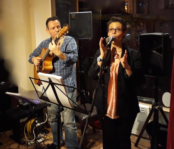 Concert en intérieur : Marina Graf chante au micro avec intensité, vêtue d’un chemisier satiné rose et d’une veste noire, aux côtés d’Eric Gombart jouant de la guitare acoustique. Derrière eux, une fenêtre laisse apparaître des lumières de nuit.