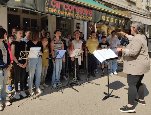 Marina Graf dirige une chorale d’adolescents et de jeunes adultes lors d’un concert en plein air devant le café culturel associatif Le Circonflexe. Les chanteurs sont regroupés autour de pupitres et de micros.
