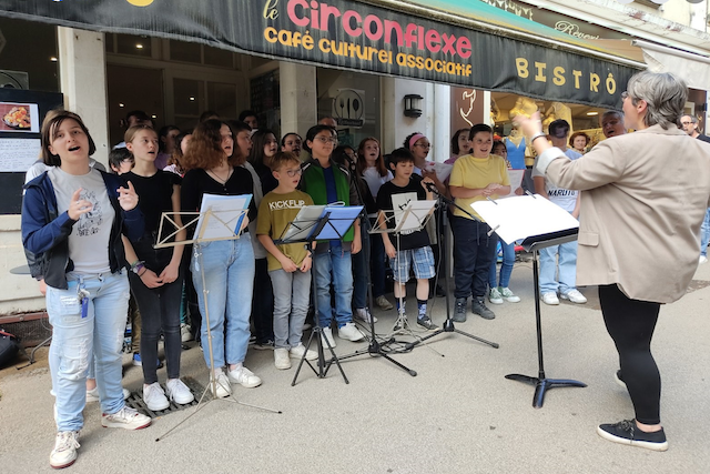 Marina Graf, de dos, dirige la même chorale devant Le Circonflexe, café culturel associatif. Les jeunes choristes chantent avec énergie, certains avec des gestes expressifs, accompagnés de pupitres à partitions et de micros.