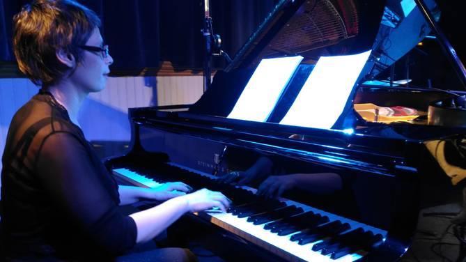 Marina Graf joue du piano à queue noir, vêtue d’un haut noir transparent, concentrée sur une partition posée devant elle. Lumière bleue créant une atmosphère intimiste de concert privé.