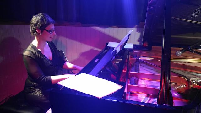 Marina Graf joue du piano à queue, habillée d’une robe noire élégante et transparente sur les bras, concentrée sur sa partition. La scène est éclairée de lumières violettes et bleues qui se reflètent sur l’instrument.