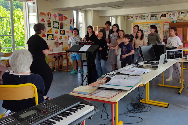 Marina Graf de dos dirige un groupe d’enfants dans une salle de classe colorée. On distingue des pupitres avec partitions et des ordinateurs portables sur les tables. Des dessins d’élèves décorent les murs.