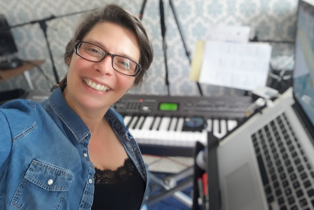 Marina Graf souriante, portant une chemise en jean et des lunettes, pose devant un clavier numérique et un ordinateur portable allumé. L’image montre un environnement de home studio avec partitions et matériel d’enregistrement en arrière-plan.
