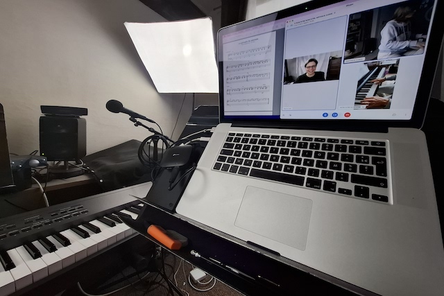 Vue rapprochée d’un poste de travail avec ordinateur portable affichant un cours de piano en visioconférence sur Skype. Marina Graf apparaît souriante à l’écran, aux côtés d’un élève jouant du piano. Un clavier et un micro sont visibles devant l’ordinateur.