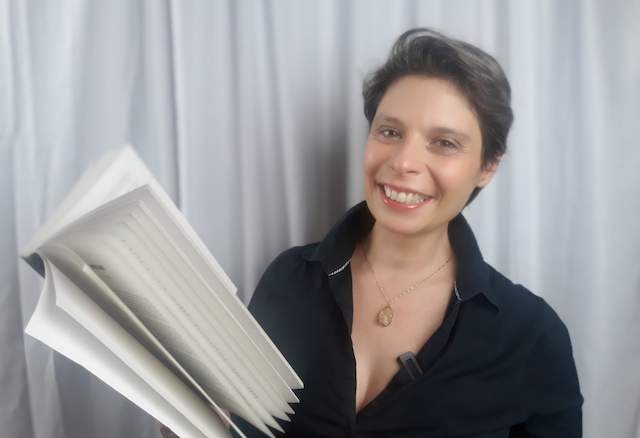Portrait de Marina Graf souriante, portant une chemise noire légèrement ouverte, un pendentif doré et tenant un livre ouvert vers la caméra. L’arrière-plan est un rideau blanc.