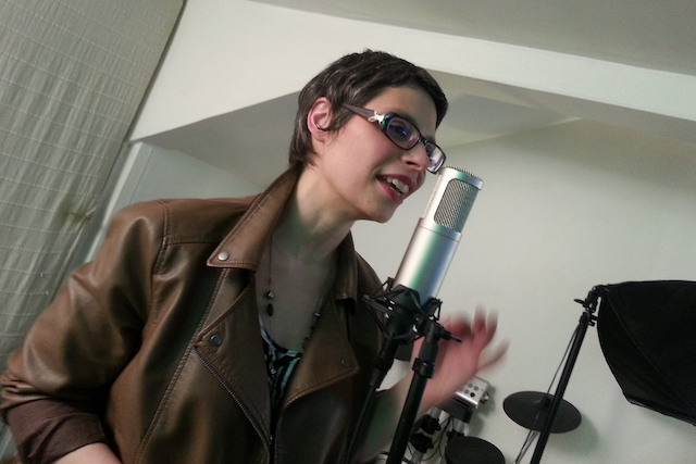 Marina Graf, portant une veste en cuir marron et des lunettes, chante debout devant un micro de studio. Elle lève légèrement la main droite en interprétant, avec en arrière-plan du matériel de musique et un rideau clair.