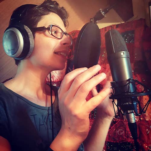 Marina Graf en studio d’enregistrement, portant un casque audio argenté et noir. Elle parle devant un grand micro équipé d’un filtre anti-pop rond. Elle sourit légèrement, avec en arrière-plan un tissu décoratif coloré rouge et orange.