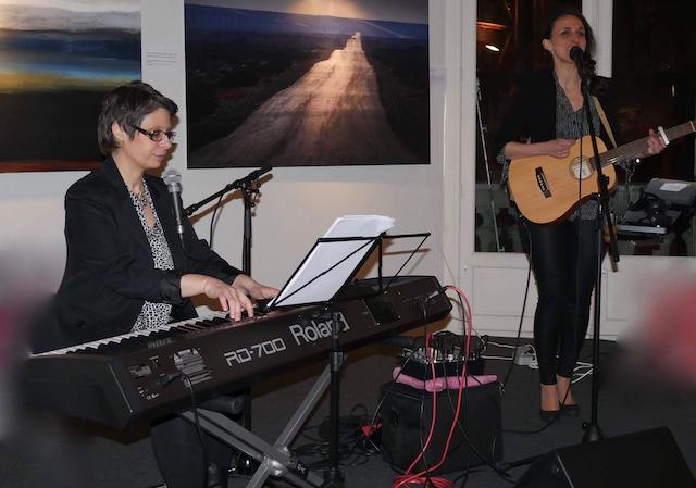 Duo musical lors d’un événement privé dans une galerie d’art. Marina Graf est assise au piano électrique, vêtue d’une veste noire et d’un haut à motifs, accompagnant Sophie Almanza, debout à la guitare acoustique et chantant au micro. En arrière-plan, des œuvres d’art sont exposées.