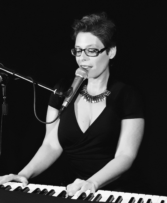 Marina Graf, chanteuse et pianiste, joue sur un clavier et chante dans un micro. Photo en noir et blanc, elle porte des lunettes, une robe noire décolletée et un collier large. L’éclairage met en valeur son visage et ses mains sur le piano.