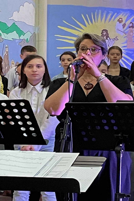 Marina Graf, chanteuse, interprète une chanson au micro sur scène. Elle est entourée d’un groupe de jeunes choristes habillés en blanc, avec une fresque colorée représentant Jésus et des rayons lumineux en arrière-plan.