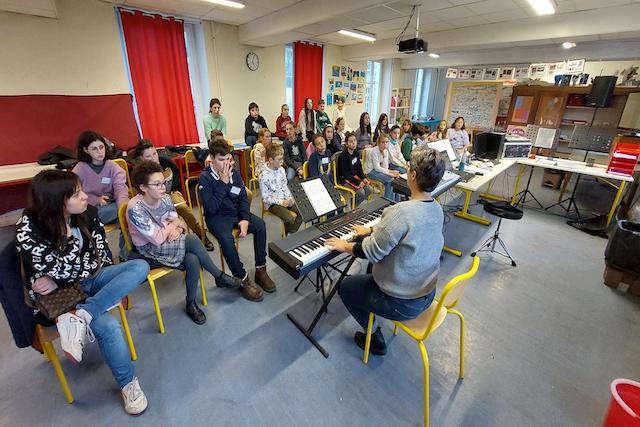 Classe d’adolescents assis sur des chaises jaunes, observant marina graf jouant du piano électrique. Marina graf, vue de dos, porte un pull gris. Salle de classe avec décor scolaire, affiches au mur et rideaux rouges. c'est la chorale Vox caeli du collège delfeuille.