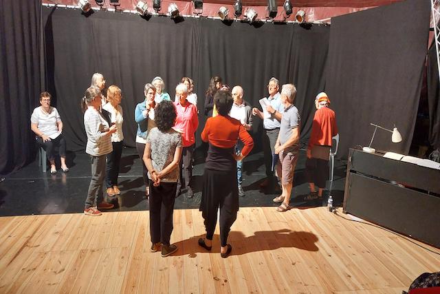 Groupe d’une quinzaine de choristes adultes sur scène, dirigés par une femme en rouge et noir vue de dos. Les choristes sont disposés en demi-cercle, certains tenant des partitions. Fond noir de théâtre avec projecteurs au-dessus. Marina Graf donne des instructions de chant