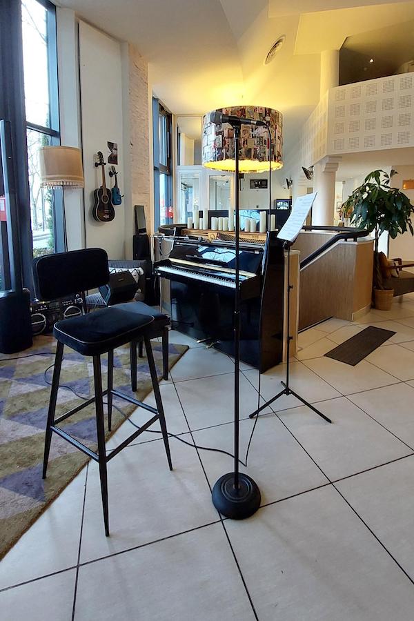 Salle moderne avec un piano droit noir, un tabouret haut et un micro sur pied. Décor chaleureux avec lampadaire recouvert de photos, guitares accrochées au mur et grande baie vitrée laissant entrer la lumière du jour.