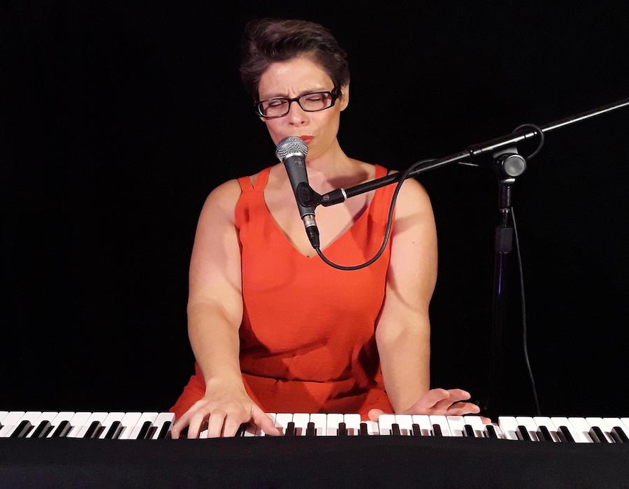Marina Graf assise au piano électrique, habillée d’une robe rouge sans manches. Elle chante dans un micro, yeux fermés et expression intense, les mains sur le clavier. Fond noir neutre.