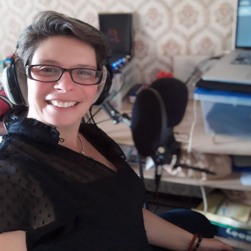 Marina Graf souriante, assise avec un casque sur les oreilles dans son espace d’enregistrement à domicile. Devant elle, un micro professionnel et un ordinateur portable sont installés sur un bureau.