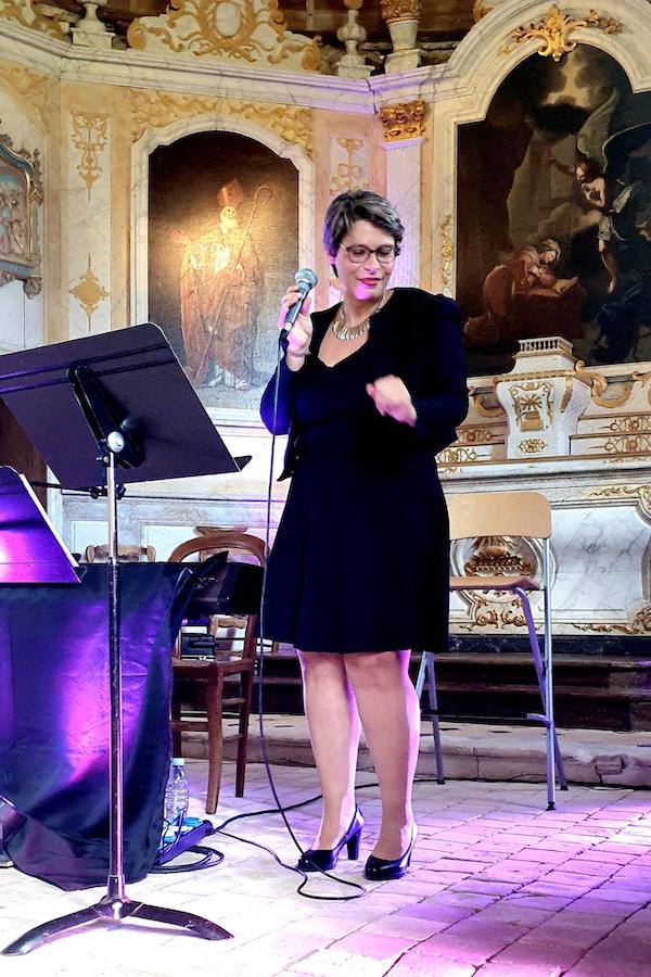 Marina Graf est debout sur scène dans une église décorée de fresques religieuses, portant une robe noire et une veste assortie, tenant un micro et semblant danser légèrement. Des projecteurs projettent une lumière violette sur le sol.