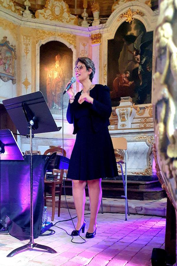 Marina Graf est debout dans la même église baroque, robe et veste noires, tenant un micro à la main et chantant avec énergie. Des pupitres et un piano électrique sont visibles à gauche.