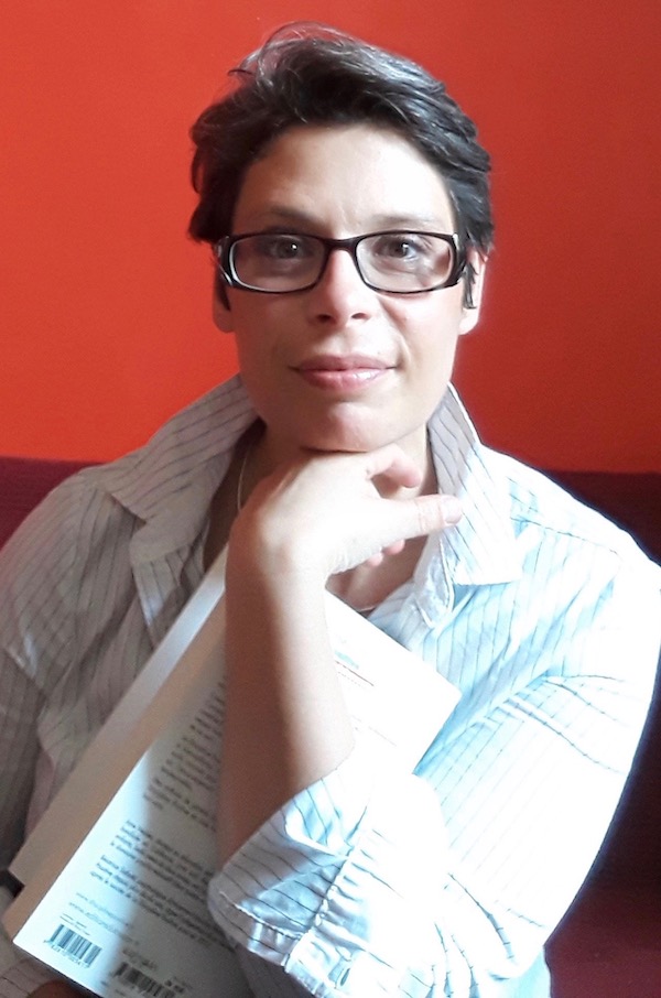 Portrait de Marina Graf assise devant un fond rouge vif. Elle porte une chemise blanche à fines rayures, des lunettes noires et tient un livre ouvert contre elle. Son menton repose sur sa main gauche, expression douce et réfléchie.