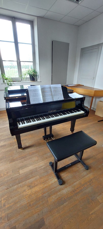 Piano à queue noir Yamaha dans une salle lumineuse, posé sur un parquet en bois. Une partition est ouverte sur le pupitre, une banquette noire est placée devant l’instrument. Une fenêtre laisse entrer la lumière du jour, avec deux plantes vertes posées sur le rebord.
