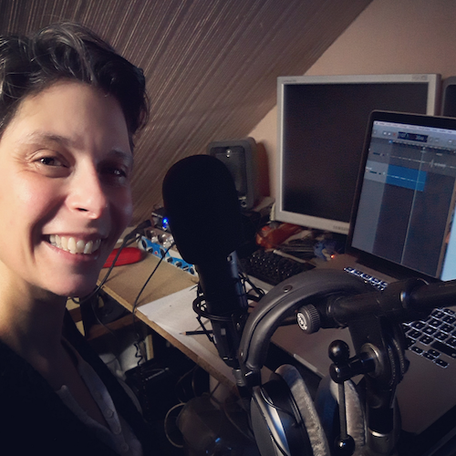 Marina Graf souriante, assise dans son home studio, portant un casque audio et des lunettes, devant un micro et un ordinateur portable affichant un logiciel d’enregistrement audio. Des câbles et du matériel de sonorisation entourent le bureau.