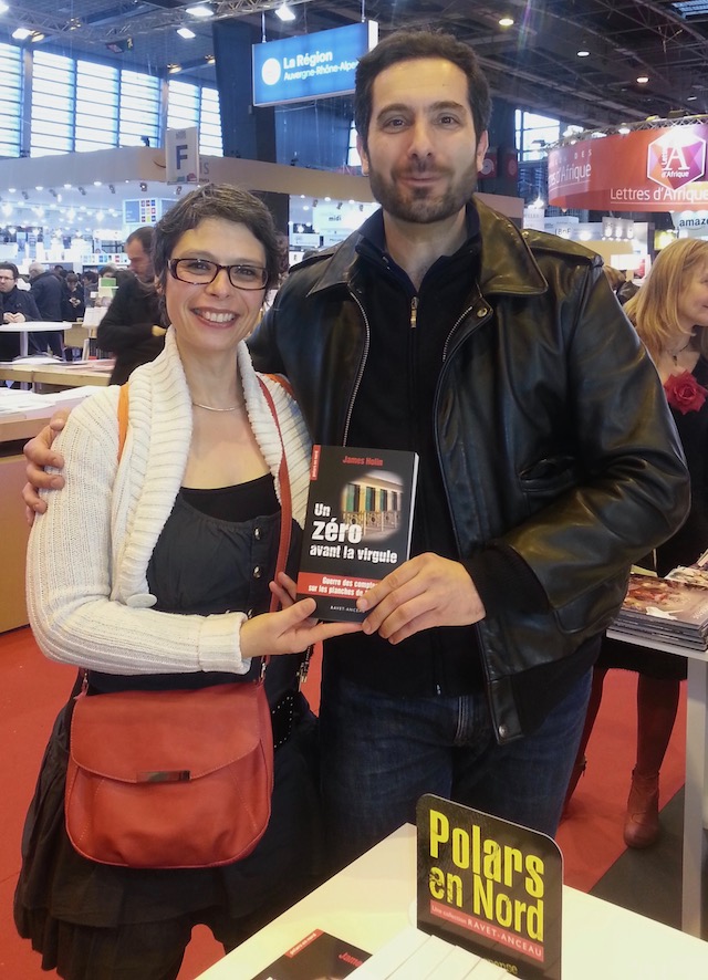 Photo prise au Salon du Livre de Paris. Marina Graf sourit aux côtés d’un homme en blouson de cuir noir qui s'appelle James Holin, qui tient son livre intitulé Un zéro avant la virgule. Elle porte un gilet blanc, un haut noir et un sac rouge en bandoulière.