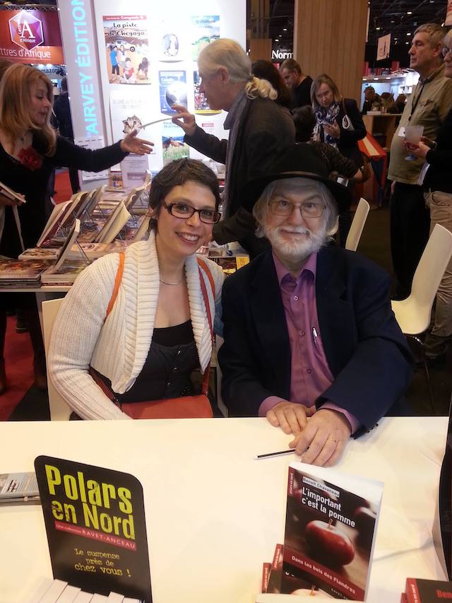 Marina Graf assise à une table du Salon du Livre de Paris aux côtés de l’auteur Benoît Chavaneau, qui porte un chapeau noir et une veste sombre. Devant eux, plusieurs exemplaires du livre L’important c’est la pomme. Autour, on distingue d’autres stands et visiteurs.