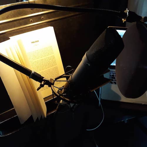 Un micro de studio professionnel en gros plan, monté sur suspension avec un filtre anti-pop rond devant. À côté, un livre ouvert sur un pupitre, éclairé par une lampe de lecture. Un ordinateur portable est visible en arrière-plan.