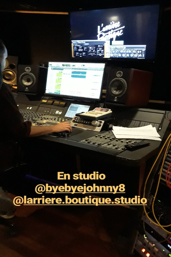 Vue d’un studio d’enregistrement professionnel avec une grande console de mixage, des écrans affichant un logiciel audio, et des enceintes de monitoring. Un ingénieur du son travaille devant la console. Texte incrusté : « En studio @byebyejohnny8 @larriere.boutique.studio