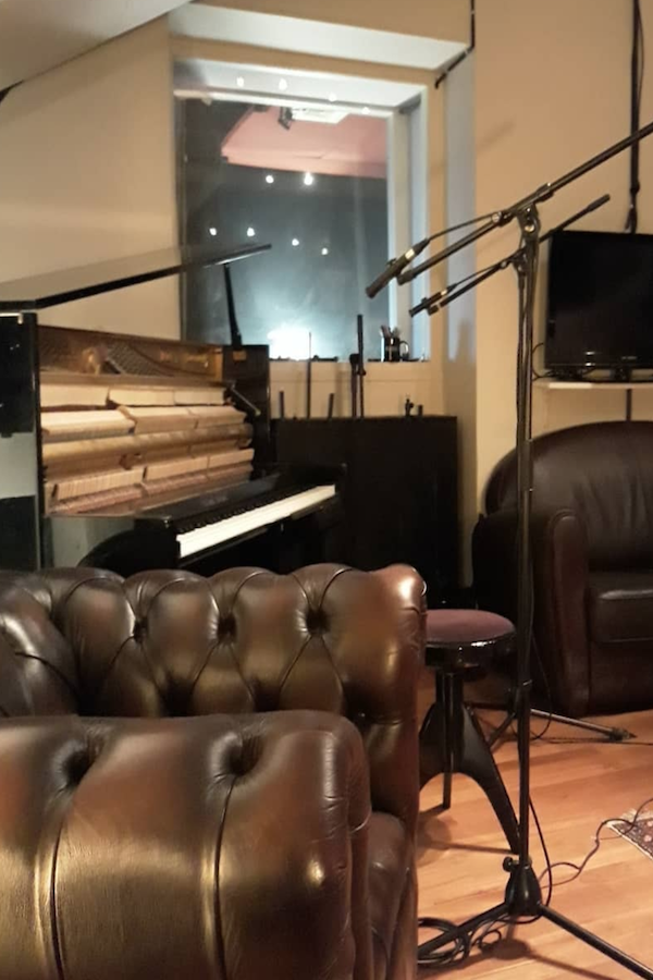 Intérieur d’un studio d’enregistrement avec un piano à queue ouvert, des micros sur pied, des fauteuils en cuir et une vitre donnant sur la cabine d’enregistrement. Atmosphère professionnelle et chaleureuse.