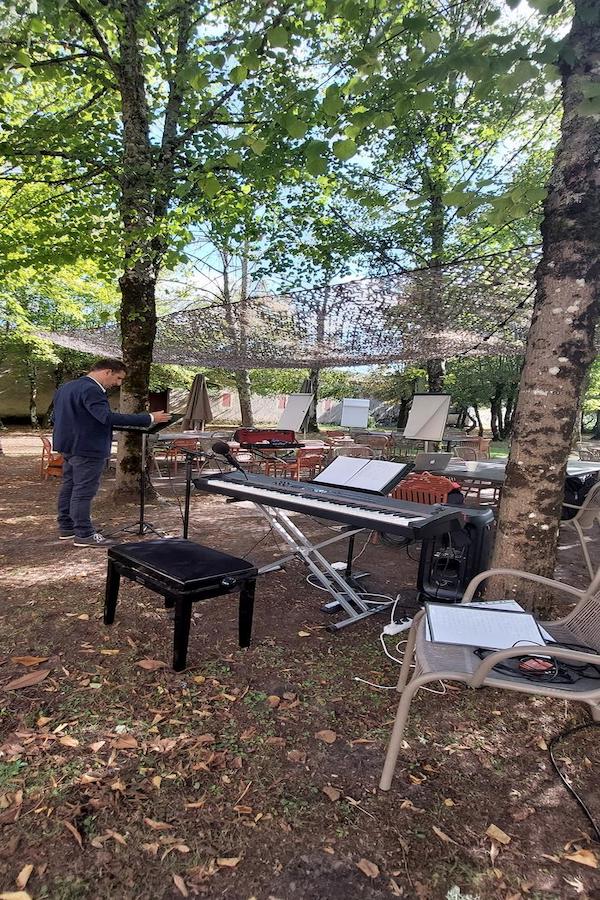 Un espace extérieur ombragé sous les arbres, aménagé avec un piano numérique sur pied, des pupitres et des chaises disposées pour les participants. L’ambiance est prête pour accueillir un atelier de team building musical dirigé par Marina Graf, qui va bientôt commencer. Un homme en costume prépare un pupitre, tandis que le matériel (micro, câbles, partitions, ordinateur portable) est déjà installé pour l’animation.