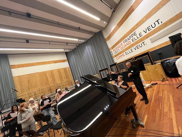 Salle de répétition moderne aux studios de La Seine Musicale, avec un grand piano à queue noir au centre. Marina Graf est assise au piano, en train d’animer une séance musicale avec un groupe de participants debout tenant des feuilles de chant. Un coach en noir se tient à côté d’elle. Sur le mur blanc est inscrite une citation en grandes lettres : « La musique frappe où elle veut, quand elle veut, comme elle veut.