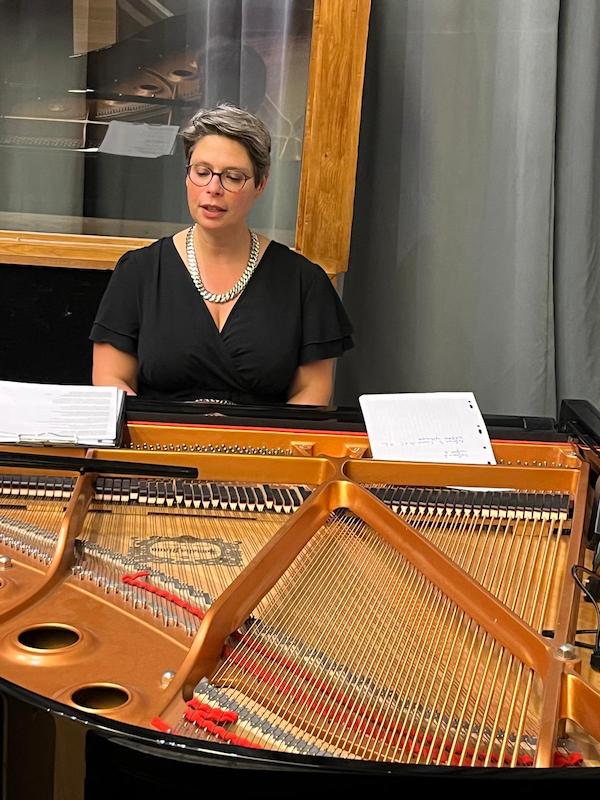 Plan rapproché de Marina Graf assise derrière un piano à queue ouvert, partitions posées devant elle. Elle porte une robe noire avec un collier argenté et des lunettes, concentrée dans son animation musicale. Derrière elle, un rideau gris et des panneaux acoustiques en bois.