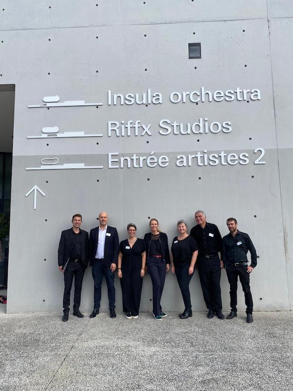 Photo de groupe prise à l’extérieur devant le mur gris clair des studios de La Seine Musicale, sur lequel on lit les inscriptions « Insula orchestra », « Riffx Studios », « Entrée artistes 2 ». Marina Graf se tient au centre, souriante, entourée de six autres coachs en tenue professionnelle noire.
