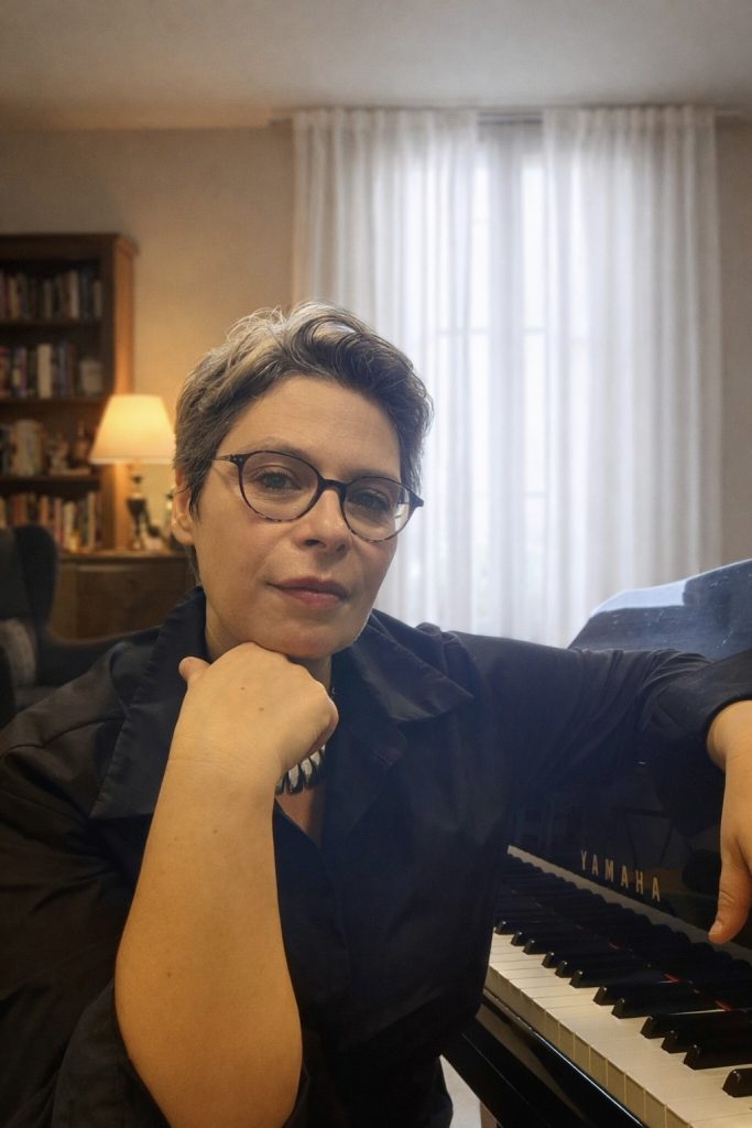 Portrait rapproché de Marina Graf, appuyée sur un piano Yamaha dans un salon lumineux. Elle porte des lunettes et une veste sombre, le menton posé sur la main, avec une expression réfléchie et posée, suggérant un moment de réflexion musicale ou pédagogique.