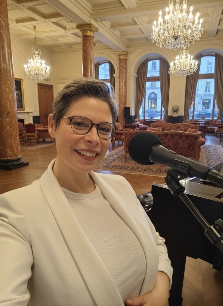 Portrait de Marina Graf, musicienne et chanteuse, souriante devant un microphone monté sur pied, dans un grand salon élégant aux lustres en cristal, colonnes en marbre et fauteuils rouges. L’espace évoque un concert privé ou une prestation musicale dans un lieu prestigieux
