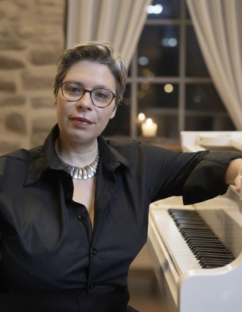 Marina Graf est assise à côté d’un piano blanc, dans une pièce chaleureuse éclairée par des bougies. Elle porte une chemise noire et un collier argenté, regarde l’objectif avec un air calme et concentré, dans une ambiance musicale intime et feutrée.
