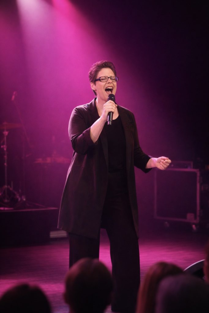 Marina Graf, chanteuse, est seule sur scène en plein concert. Elle tient un microphone à la main et chante avec intensité, la bouche grande ouverte et les yeux fermés, exprimant une forte émotion. Elle porte une tenue sombre élégante. La scène est éclairée par des lumières violettes et roses qui enveloppent l’espace, tandis que le public apparaît flou au premier plan. L’ambiance évoque une performance vocale puissante et engagée, en concert live.